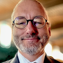 Smiling man with glasses and a beard wearing a suit, standing indoors with blurred background.
