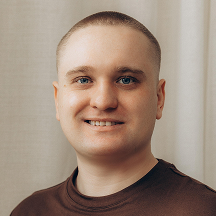 A person with short hair smiling at the camera, wearing a brown shirt, standing in front of a light-colored curtain.