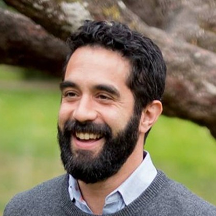 Man with dark hair and beard, smiling outdoors with a blurred natural background.