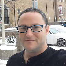A person wearing glasses and a black shirt stands outside in a snowy environment, with a white car and a brick building in the background.