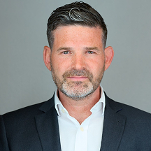 Middle-aged man with short, dark hair and a trimmed beard in a suit and white shirt against a plain gray background.
