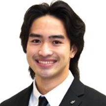 Smiling person with long dark hair, wearing a black suit and tie, against a plain white background.