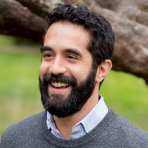 A bearded man smiling outdoors, wearing a gray sweater over a collared shirt, with blurred greenery in the background.