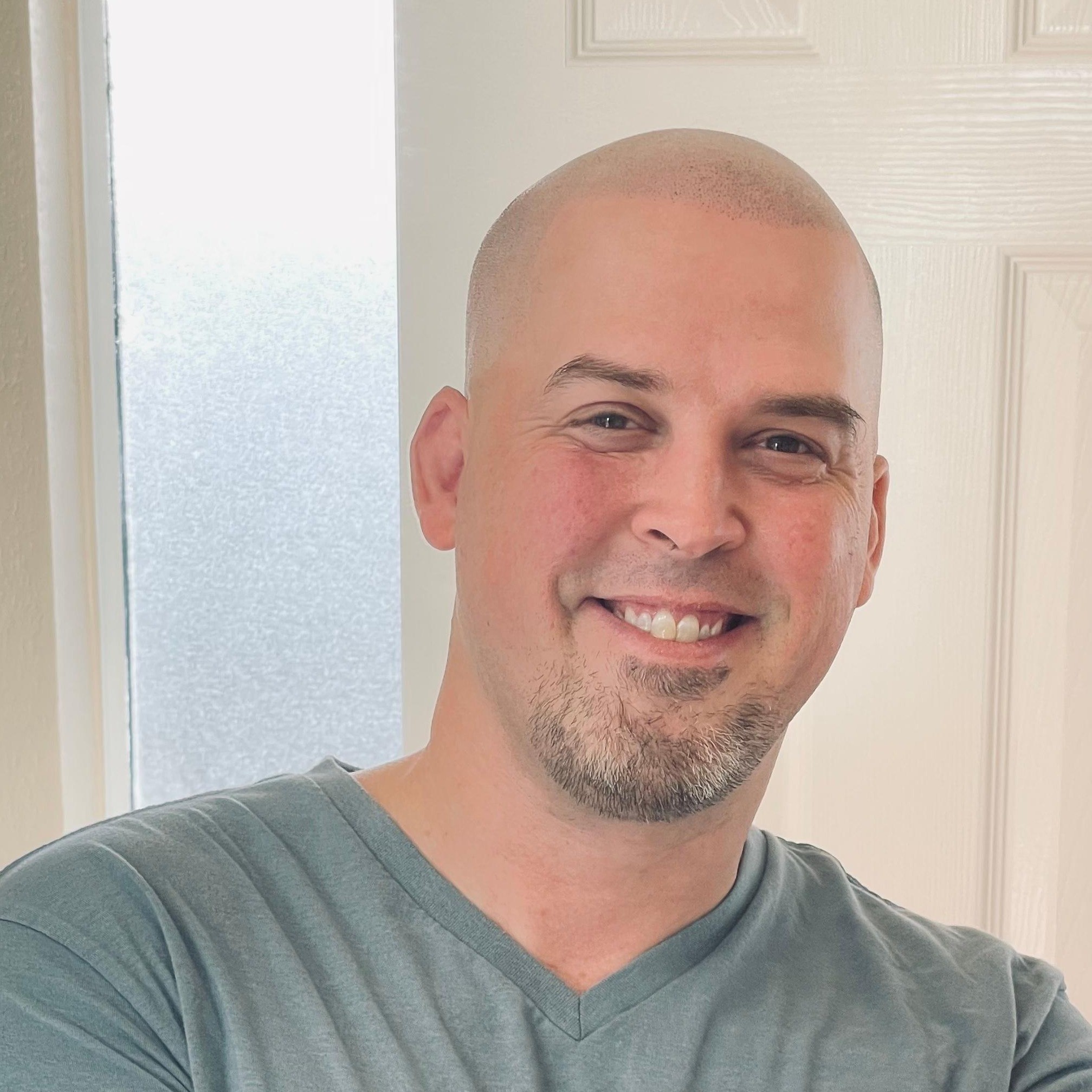 Smiling man with a shaved head and goatee wearing a light blue V-neck T-shirt, standing in a room with a white door and window.