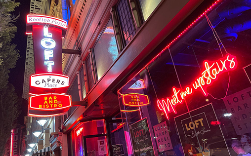 Neon signs outside a bar and bistro with words like "Rooftop Patio," "Bar and Bistro," and "Meet me upstairs" against an urban backdrop.