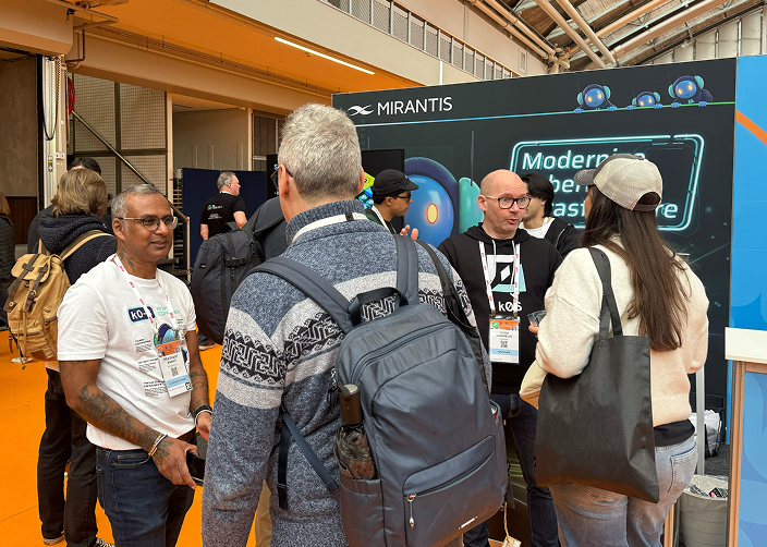 People at a tech conference booth, engaged in conversation, with the Mirantis logo and a display screen in the background.