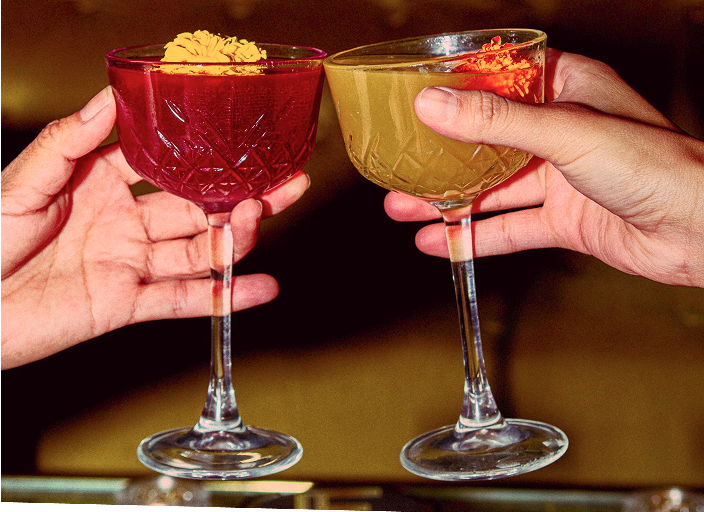 Two hands holding ornate cocktail glasses filled with colorful drinks, garnished with orange flowers, against a warm, blurred background.