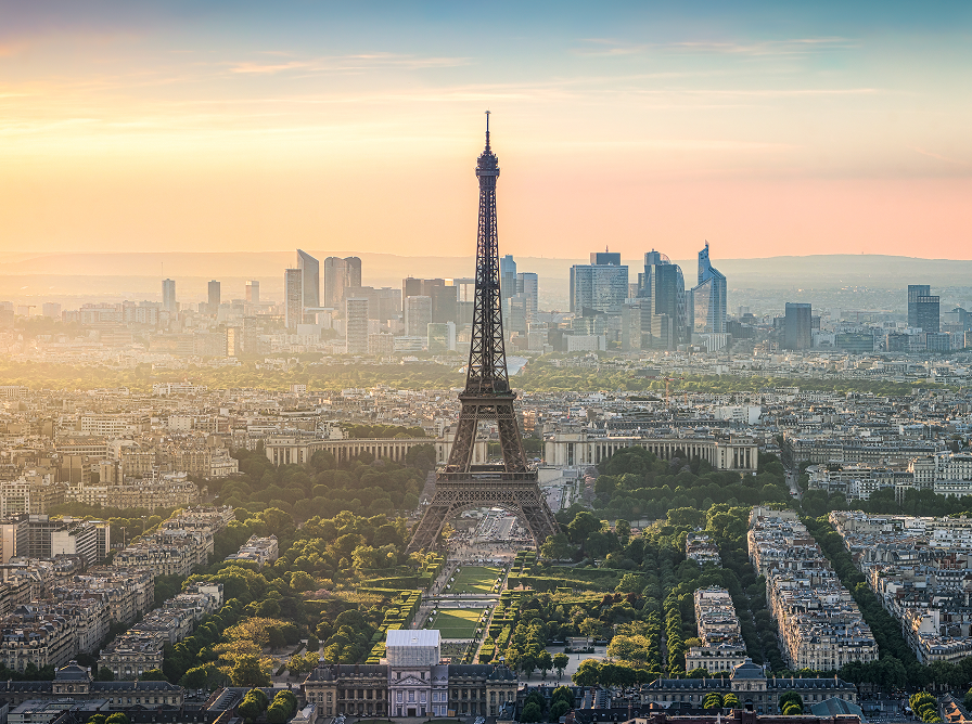 Aerial view of the Eiffel Tower at sunset, surrounded by Parisian buildings and greenery, with a modern skyline in the background.