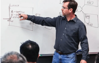 Man in a gray shirt pointing at a network diagram on a whiteboard during a lecture. Audience members visible in the foreground.