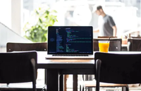 Laptop displaying code on a table with a glass of orange juice, surrounded by chairs, in a bright cafe setting.