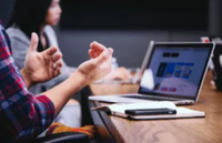 Person gesturing during a meeting, sitting at a table with a laptop, notebook, and phone. Another person is blurred in the background.