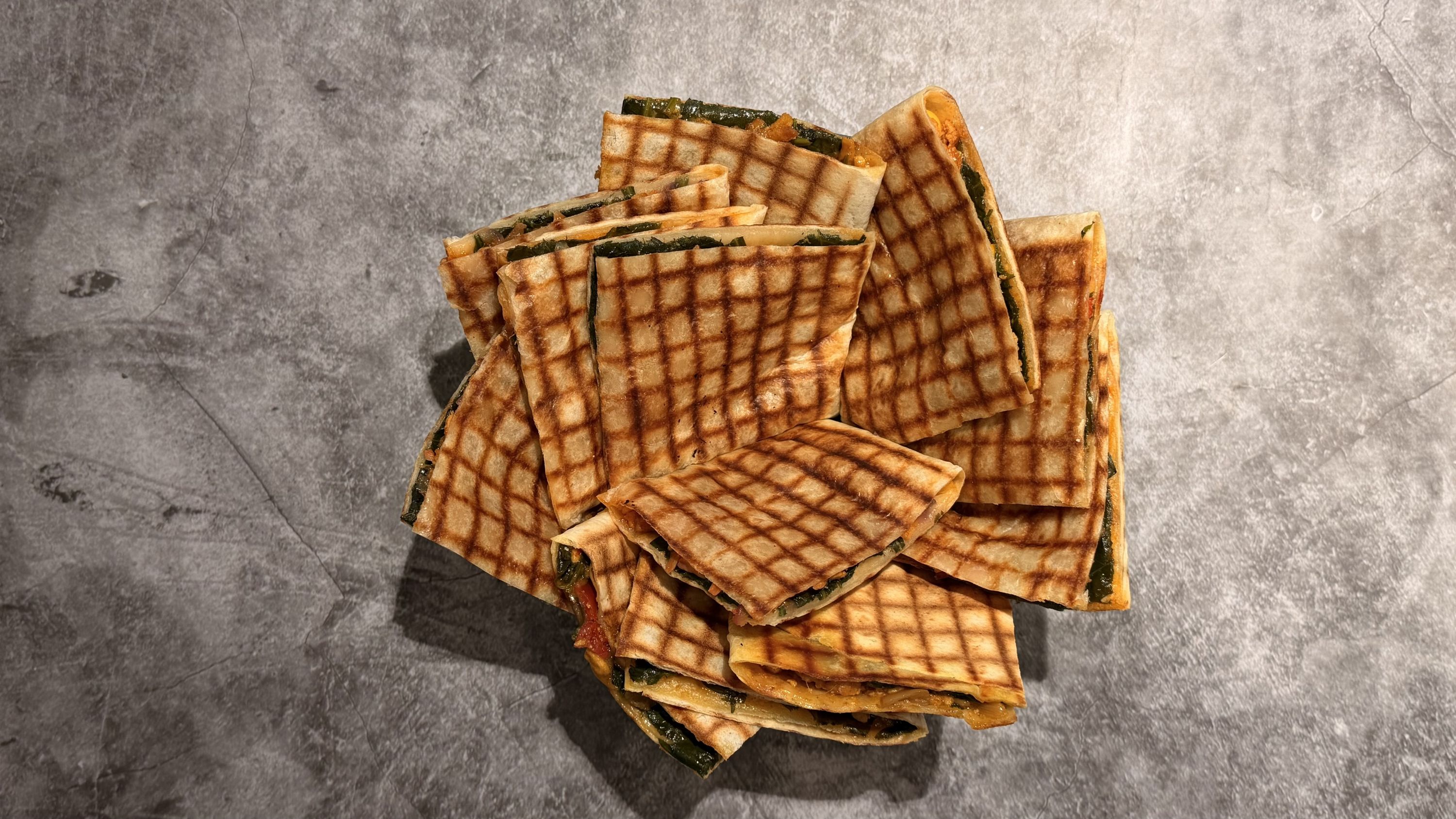 Fingerfood. A stack of grilled, folded tortillas filled with spinach and filling, arranged on a textured gray surface.