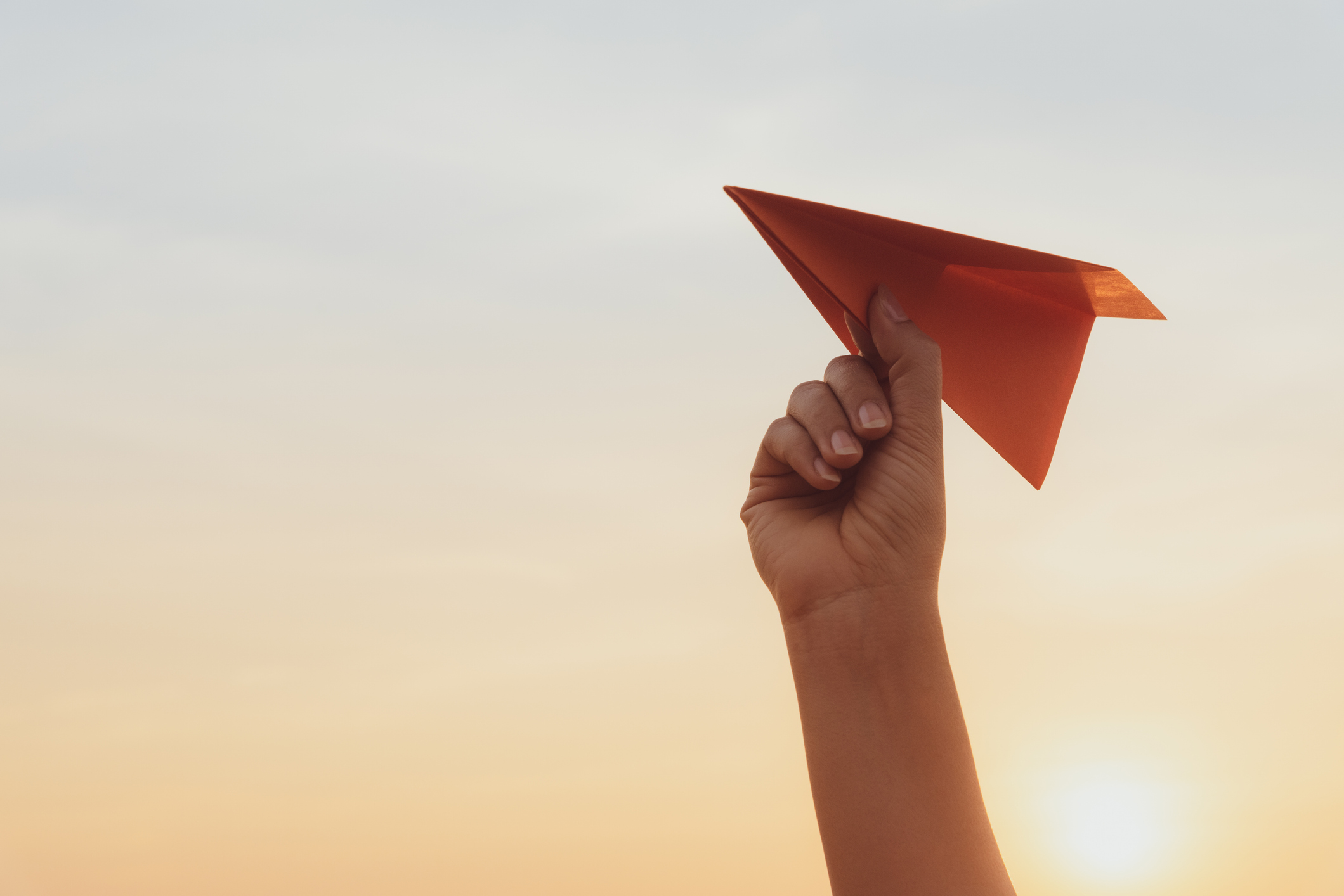 Ein Hand hält ein orangenes Papier-Flugzeug in der Hand, vor dunstigem Abendhimmel mit untergehender Sonne.