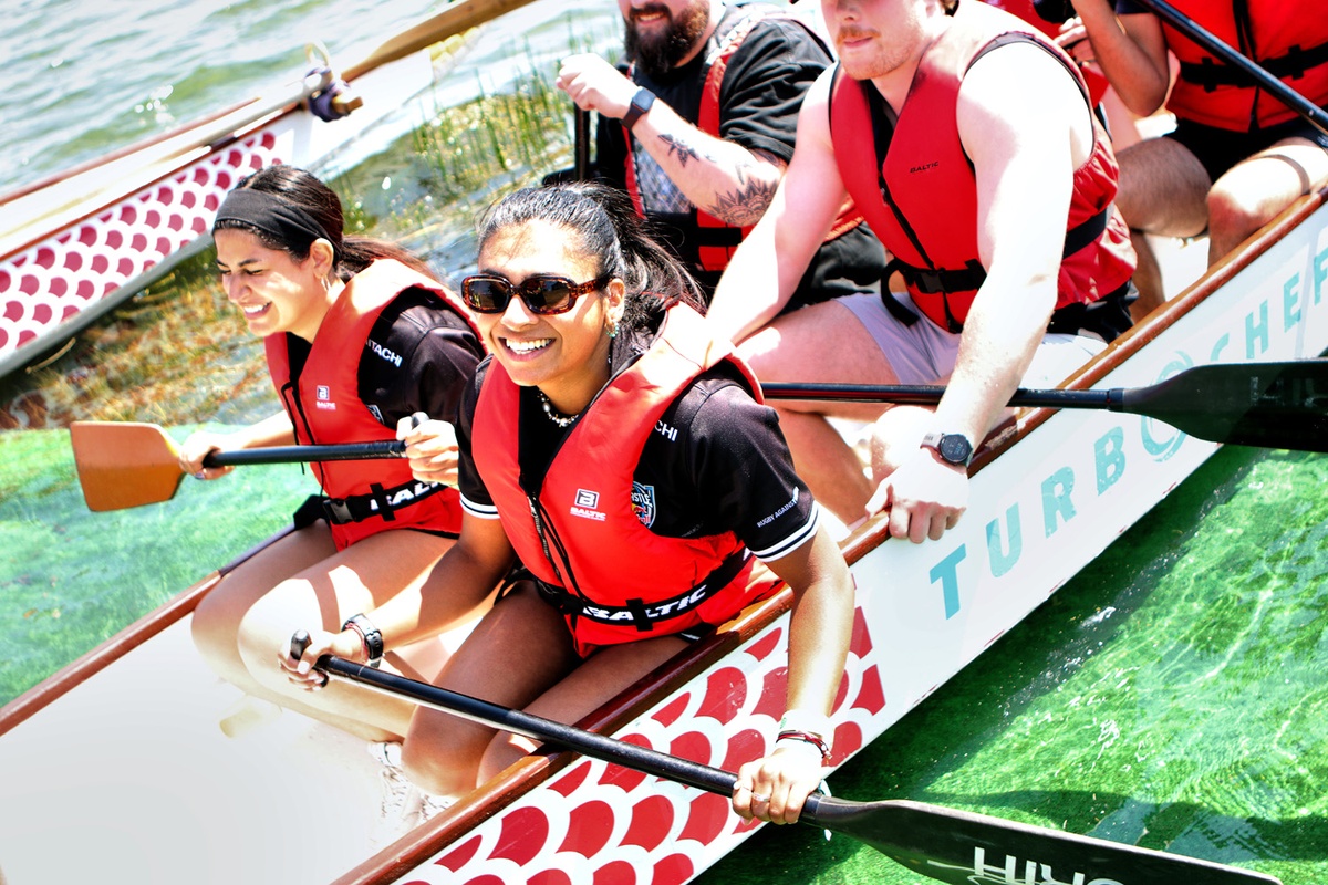 People paddling a dragon boat on the water, wearing life vests and smiling brightly in the sun.