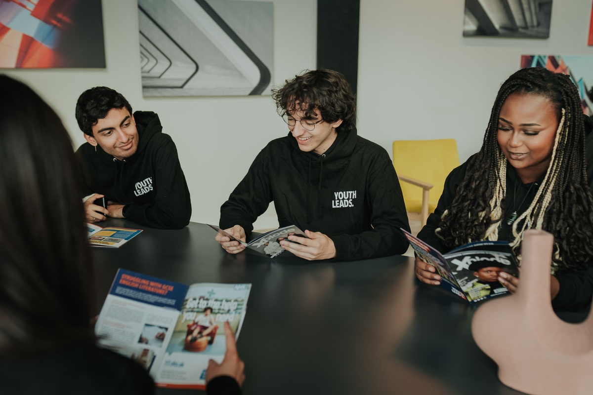 Four young adults in hoodies sit at a table reading magazines, with abstract art on the wall behind them.