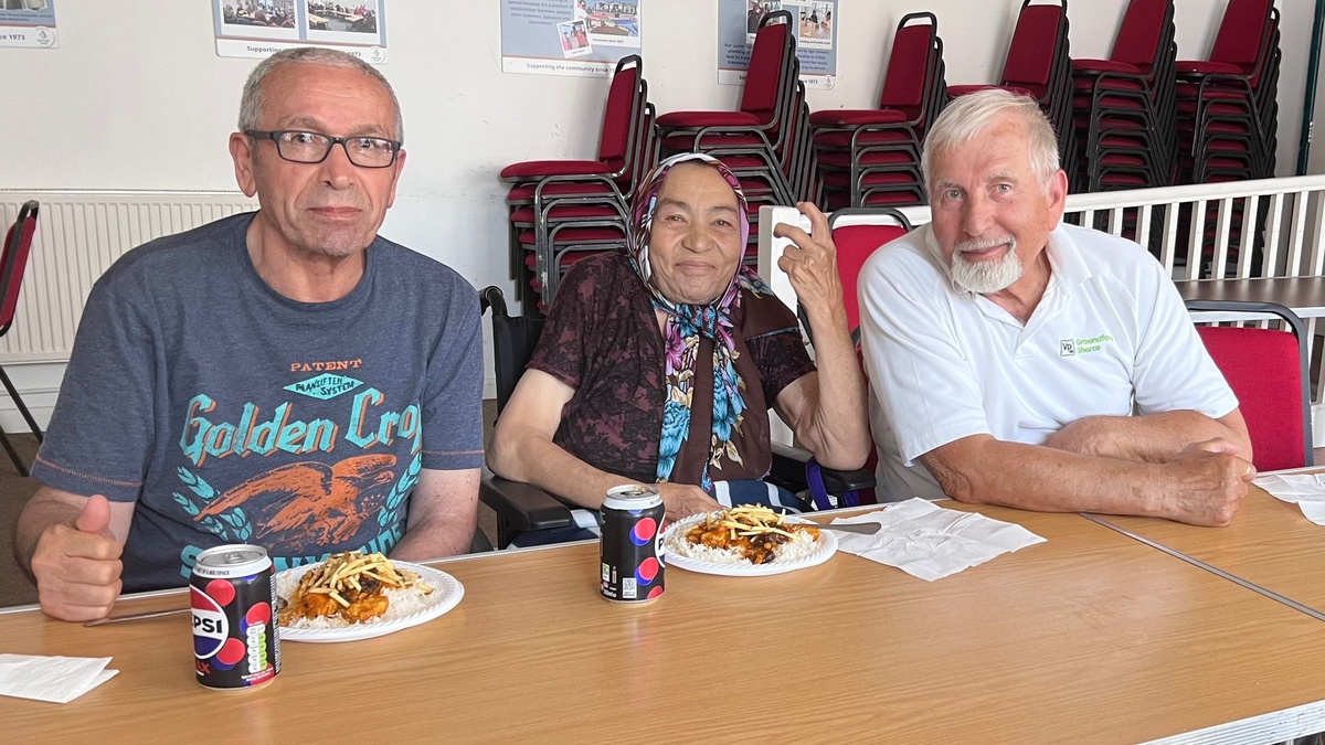 2 men sit either side of a woman wearing a headscarf as they eat a meal