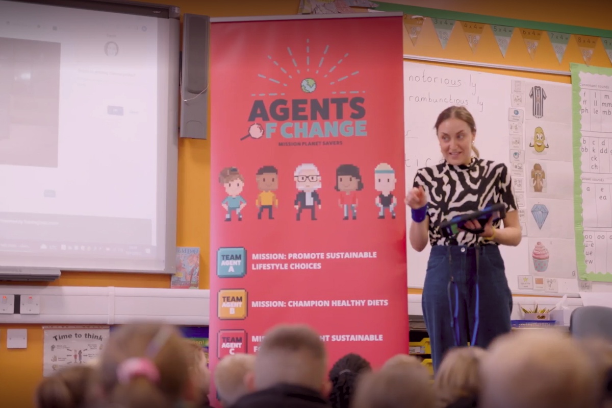 A school teacher holding a tablet standing beside a red banner with the words "Agents of change"