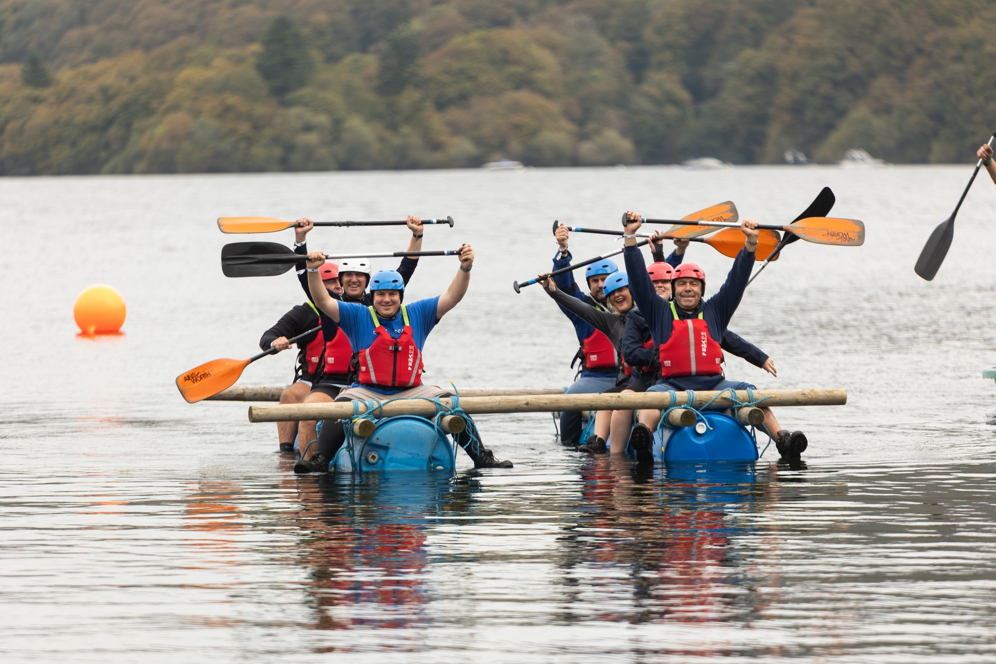 A group of people in helmets and life jackets paddle a makeshift raft on a lake, celebrating with raised paddles.