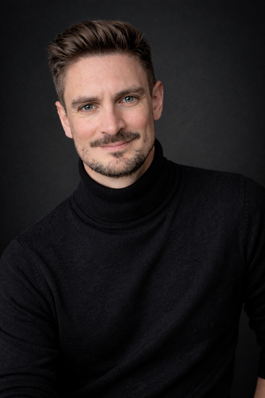 A man with short brown hair and a beard, wearing a black turtleneck, smiles warmly against a dark background.