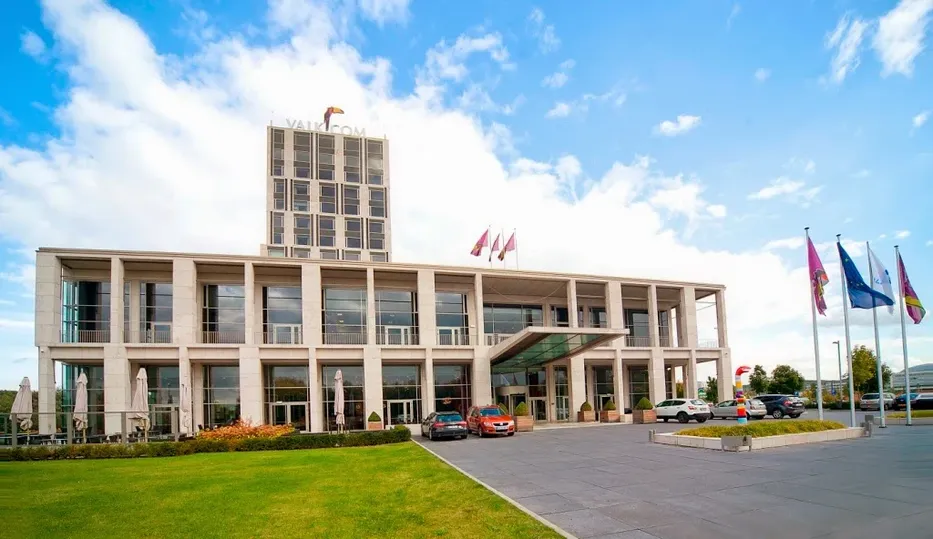 Modern hotel with large windows and a glass entrance, surrounded by flags and parked cars, under a partly cloudy sky.