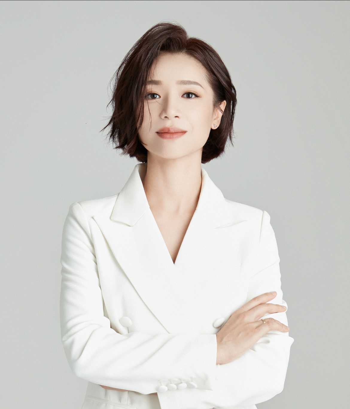 A woman with short hair wearing a white blazer, arms crossed, smiling softly against a neutral background.