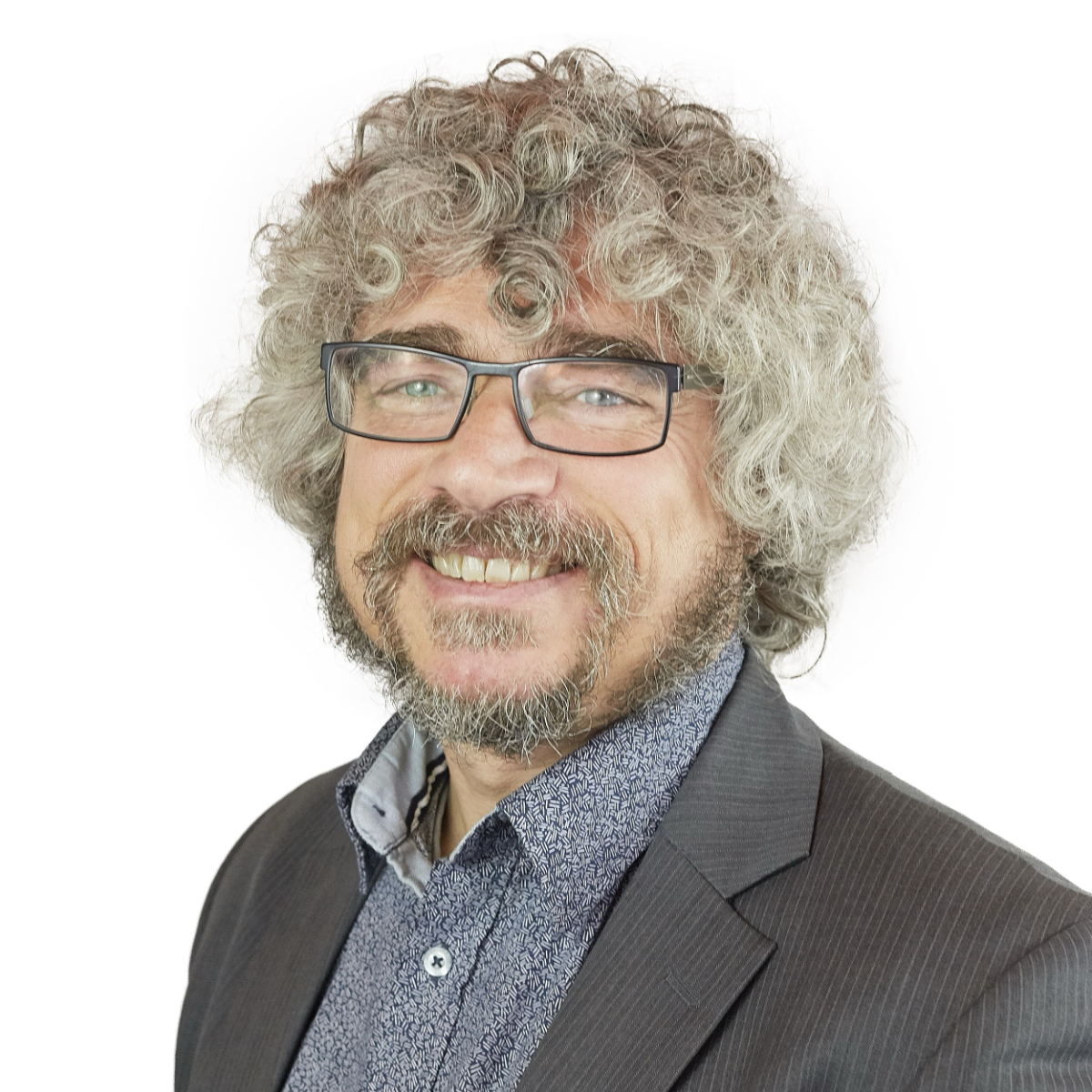 Smiling man with curly hair, glasses, and a beard wearing a suit jacket and patterned shirt, on a white background.