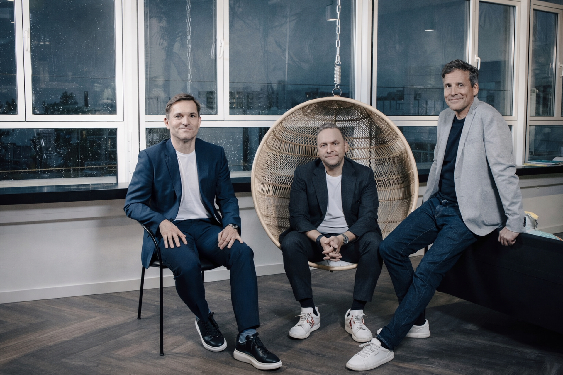 Three men in business casual attire sitting in an office; one in a hanging chair. Large windows in the background show a cityscape.