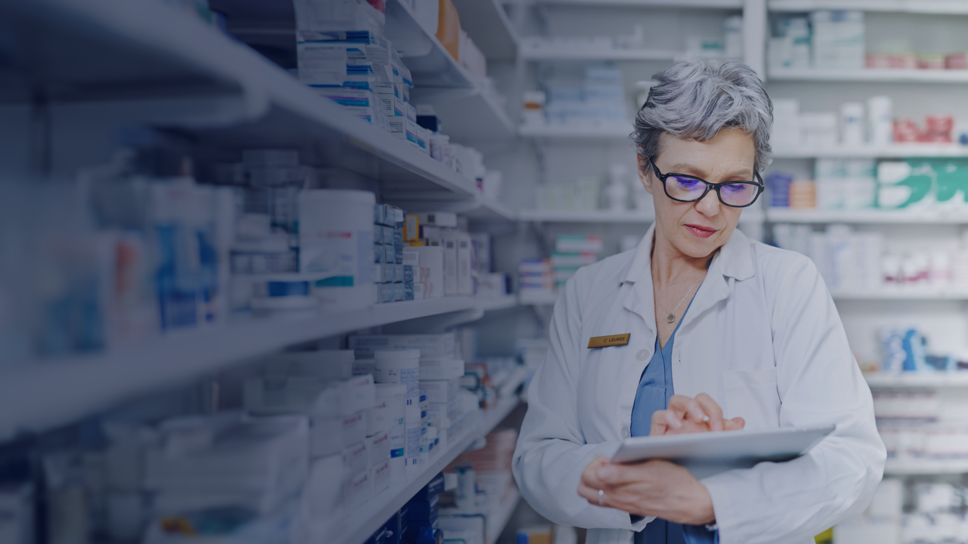 Pharmacist in a white coat and glasses reviews a tablet in a pharmacy, surrounded by shelves stocked with various medications.