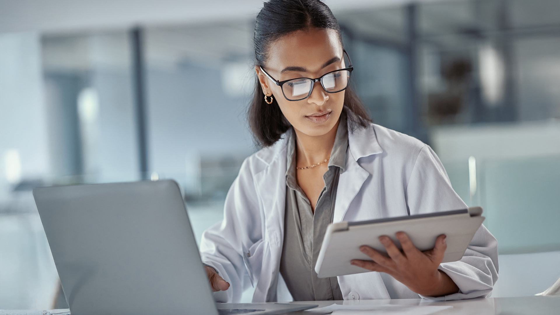 A woman in a lab coat works at a desk with a laptop and tablet in a modern office setting.