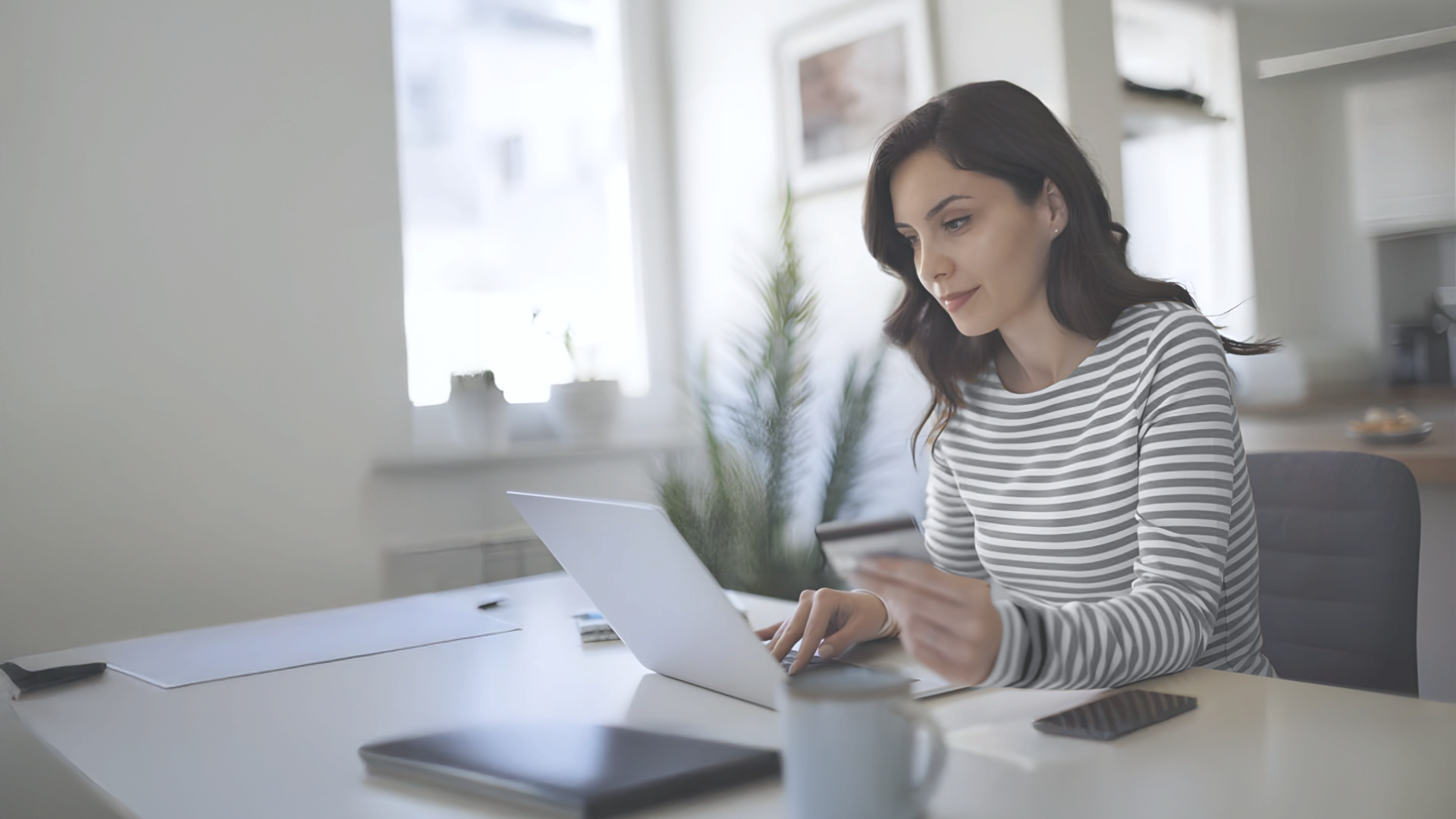 Woman in a striped shirt using a laptop at a bright, modern workspace with a smartphone and coffee mug nearby.