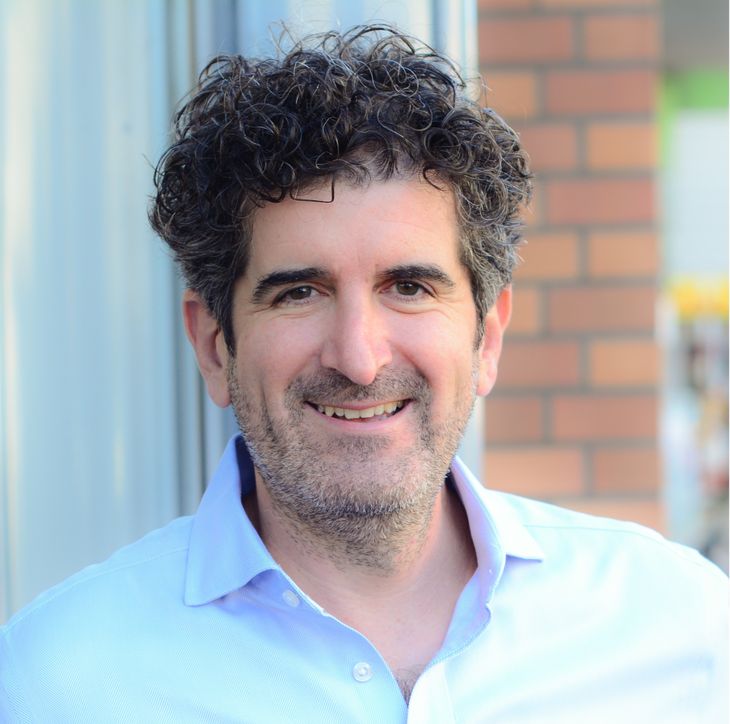 Man with curly hair smiling, wearing a light blue shirt, standing against a brick and metal background.