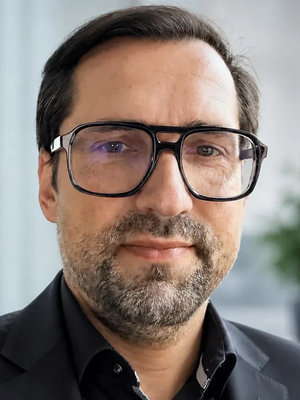Man with glasses and a beard, wearing a black suit and shirt, looks directly at the camera with a neutral expression.