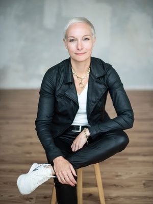 A person with short hair sits on a wooden stool, wearing a black leather jacket, white shirt, black pants, and white sneakers, in a minimalist room.