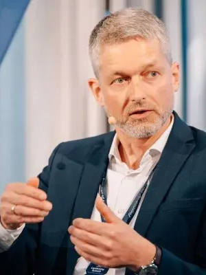 Man with short gray hair and beard wearing a suit and a microphone headset, gesturing with his hands during a talk or presentation.