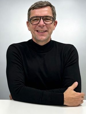 Smiling person wearing glasses and a black turtleneck, seated with arms crossed on a white surface against a neutral background.