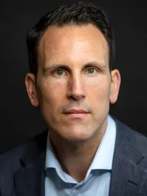 A man in a blue suit and light blue shirt poses against a dark background, looking directly at the camera with a neutral expression.
