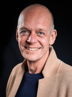 Smiling bald man in a beige blazer and dark shirt against a dark background.