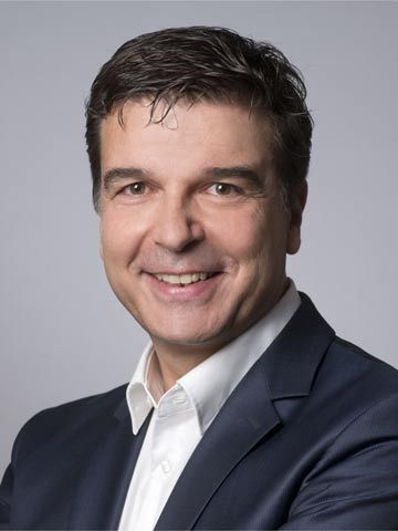 Smiling man in a suit and white shirt, with dark hair, against a gray background.