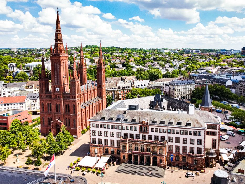 Aerial view of a historic red-brick cathedral and surrounding buildings in a vibrant cityscape under a partly cloudy sky.