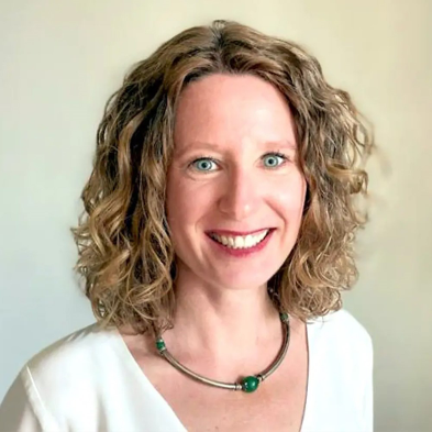 A smiling woman with curly light brown hair, wearing a white top and a necklace, against a plain background.