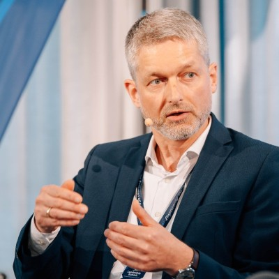 A man with short gray hair and a beard, wearing a suit and white shirt, gestures while speaking into a small microphone on his collar.