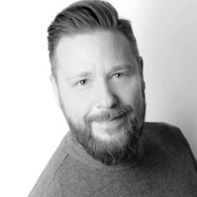Black and white portrait of a smiling man with a beard and styled hair, wearing a long-sleeve top, posing against a light background.