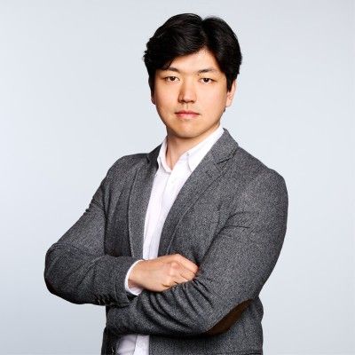 Man in a gray blazer and white shirt stands with arms crossed, looking confidently at the camera against a plain background.