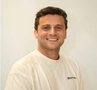 Smiling man with short dark hair wearing a white shirt with "doma." written on it, against a plain background.