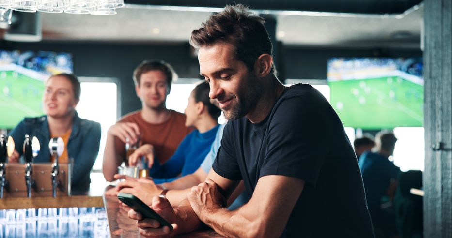 A man in a black shirt sits at a bar, smiling at his phone. People in the background watch soccer on TV screens.