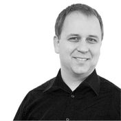 Black and white photo of a smiling individual with short hair, wearing a dark shirt, facing forward against a plain white background.
