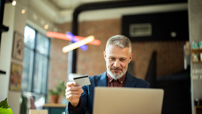 Man in a suit holding a credit card while using a laptop in a modern office with large windows and colorful decor.