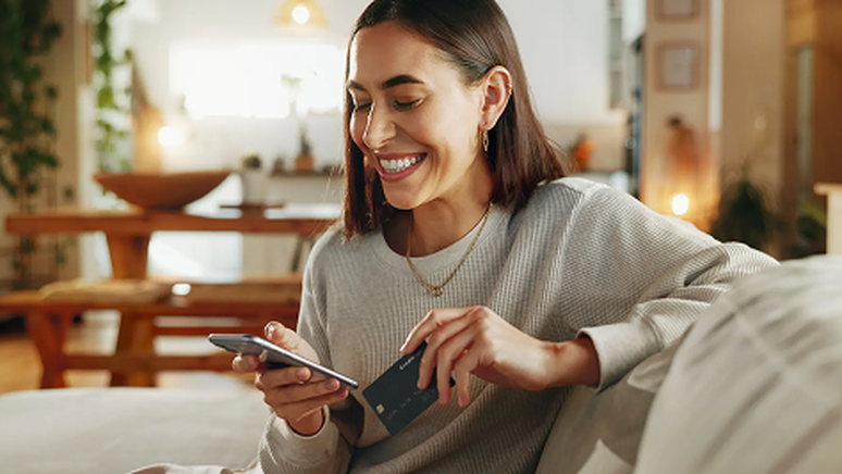 Woman smiling, sitting on a couch, holding a smartphone and a credit card in a cozy, well-lit living room.