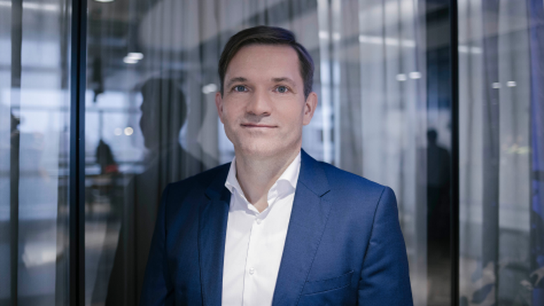 A person in a blue suit and white shirt stands indoors with glass walls and reflective surfaces in the background.