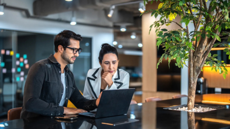 Two people in a modern office space focus on a laptop screen, surrounded by greenery and contemporary decor.